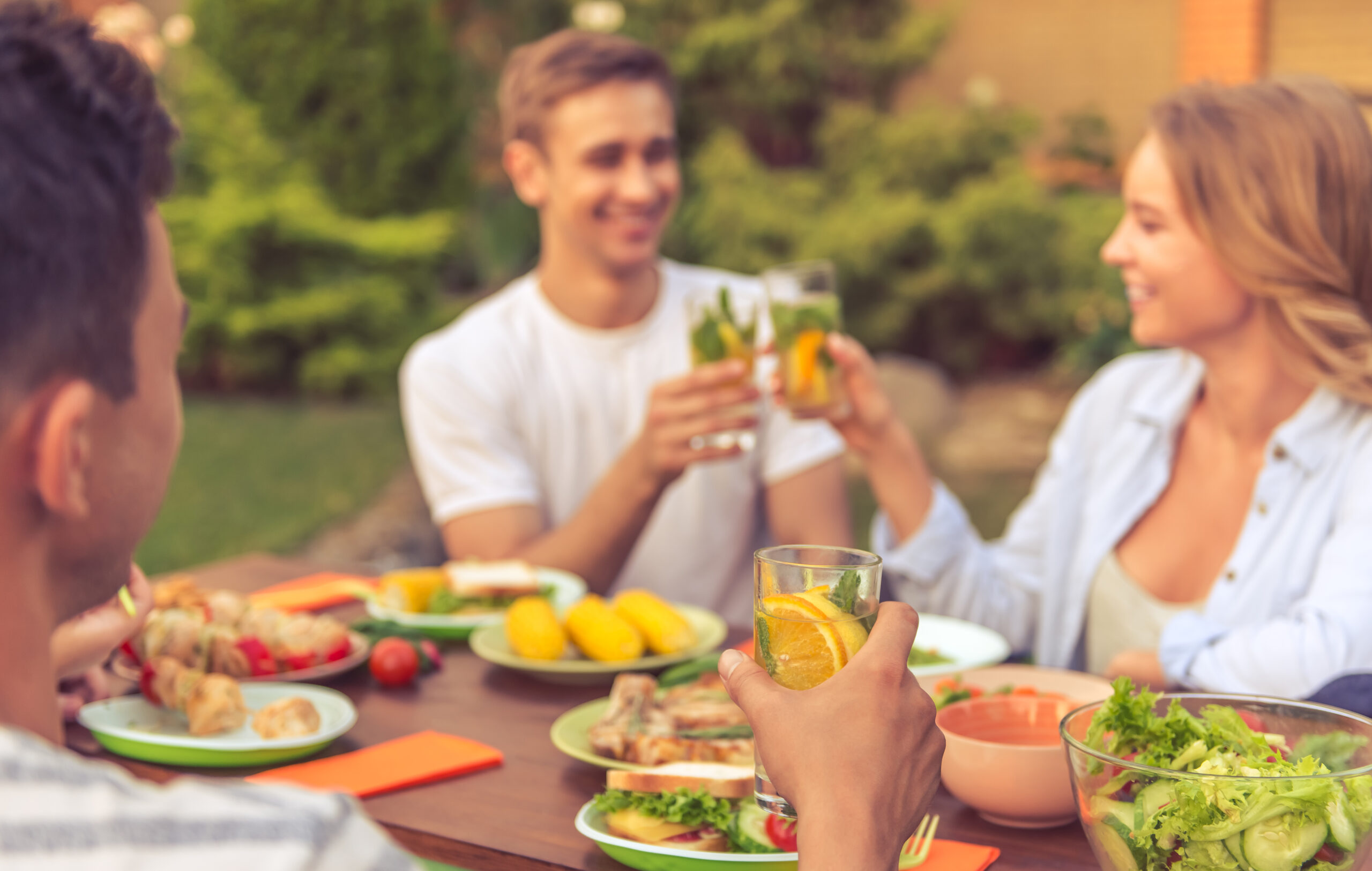 Young people are having picnic outdoors. Cropped image of young man holding a glass of lemonade while his friends are clinking glasses and smiling