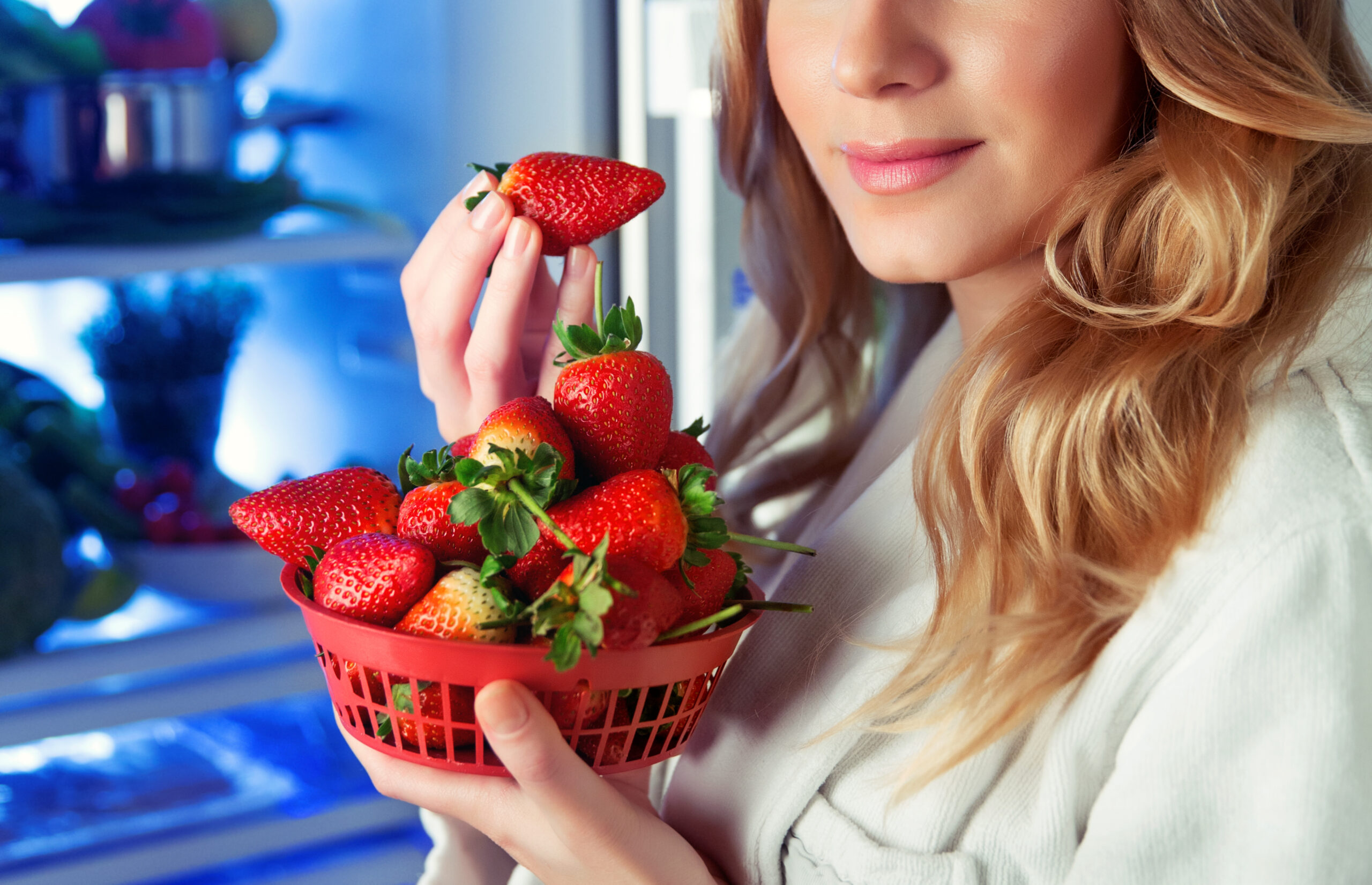 Portrait of a nice blond woman with pleasure eating fresh tasty strawberries near the open fridge, good choice, vegetarian food, weight loss, fruits diet